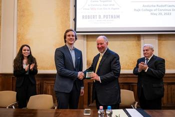 Két férfi kezet fog egy díjátadó eseményen, háttérben Robert D. Putnam előadásának címe és dátuma.