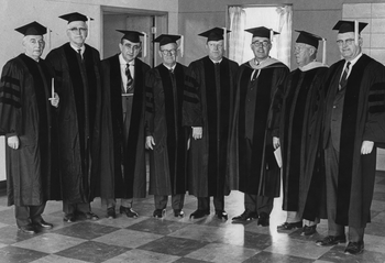 Férfiak egyetemi talárban és sapkában állnak egy szobában, diplomaosztó ünnepségen.