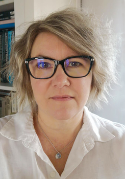 Személy fehér blúzban, nyaklánccal, könyvespolc előtt, háttérben könyvek.