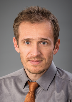 Férfi szürke inget és narancssárga nyakkendőt visel, háttér homályos.