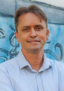 Férfi világoskék ingben áll, háttérben graffiti látható.