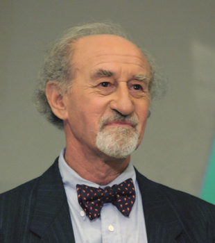 Idős férfi elegáns öltönyben és csokornyakkendőben, háttér homályos.