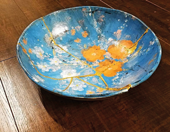 Kék alapon virágmintás kerámia tál, kintsugi technikával javítva, fa asztalon.