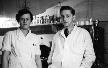 Két személy laboratóriumi köpenyben egy laboratóriumban, üvegek és eszközök a háttérben.