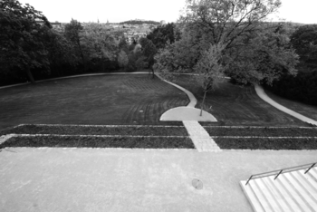 Park sétányokkal, fákkal és gondozott gyep, háttérben város panorámája.