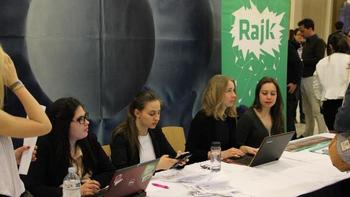 Emberek egy rendezvényen, asztalnál ülve, laptopokkal és papírokkal, háttérben Rajk banner.