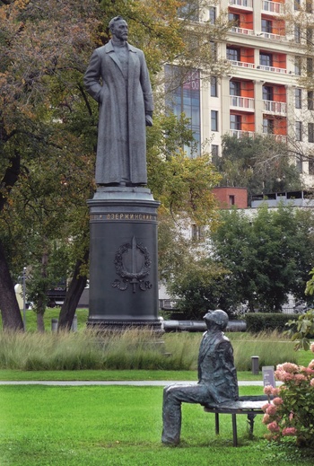 Szoborcsoport parkban, egy álló és egy ülő alak, háttérben modern épületek.