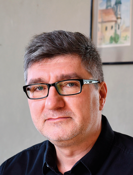 Szürke hajú férfi fekete ingben szemüveggel, háttérben festmény a falon